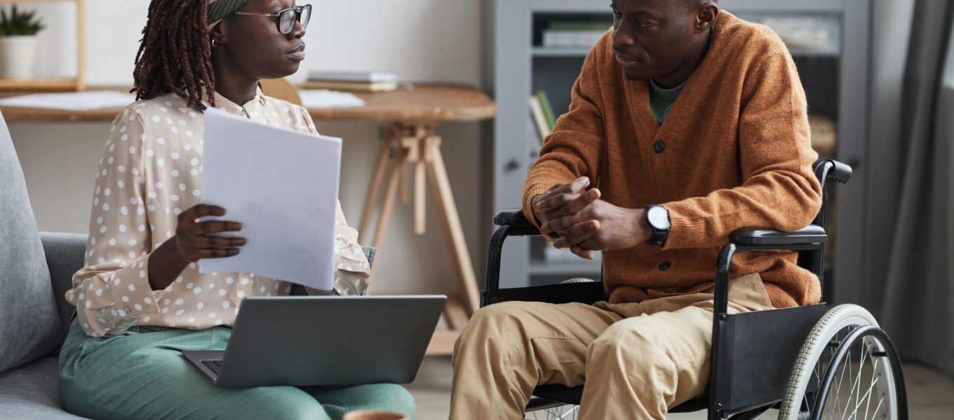 African-American Wheelchair User Talking to Business Consultant Portrait of African-American couple with handicapped man using wheelchair working from home together in modern interior