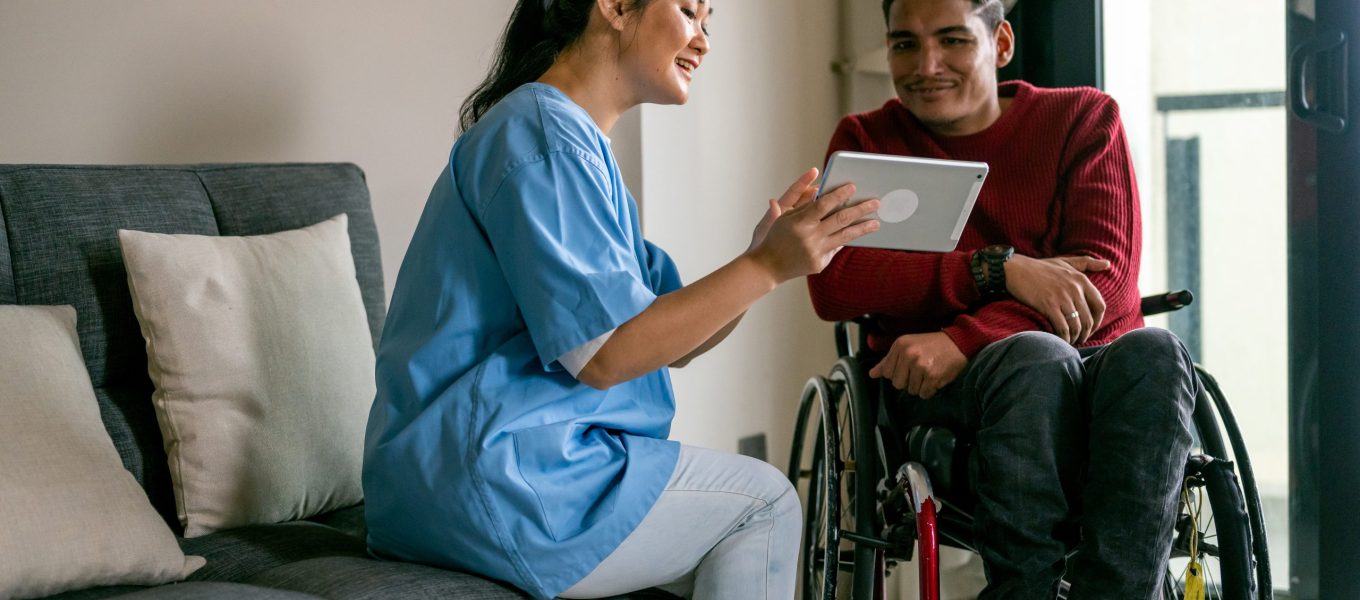 An Asian female healthcare worker teaching Mature Asian disabled Malay man in wheelchair how to online by using digital table at living room. Professional Asian female healthcare worker teaching Mature Asian disabled Malay man in wheelchair how to online by using digital table at living room.